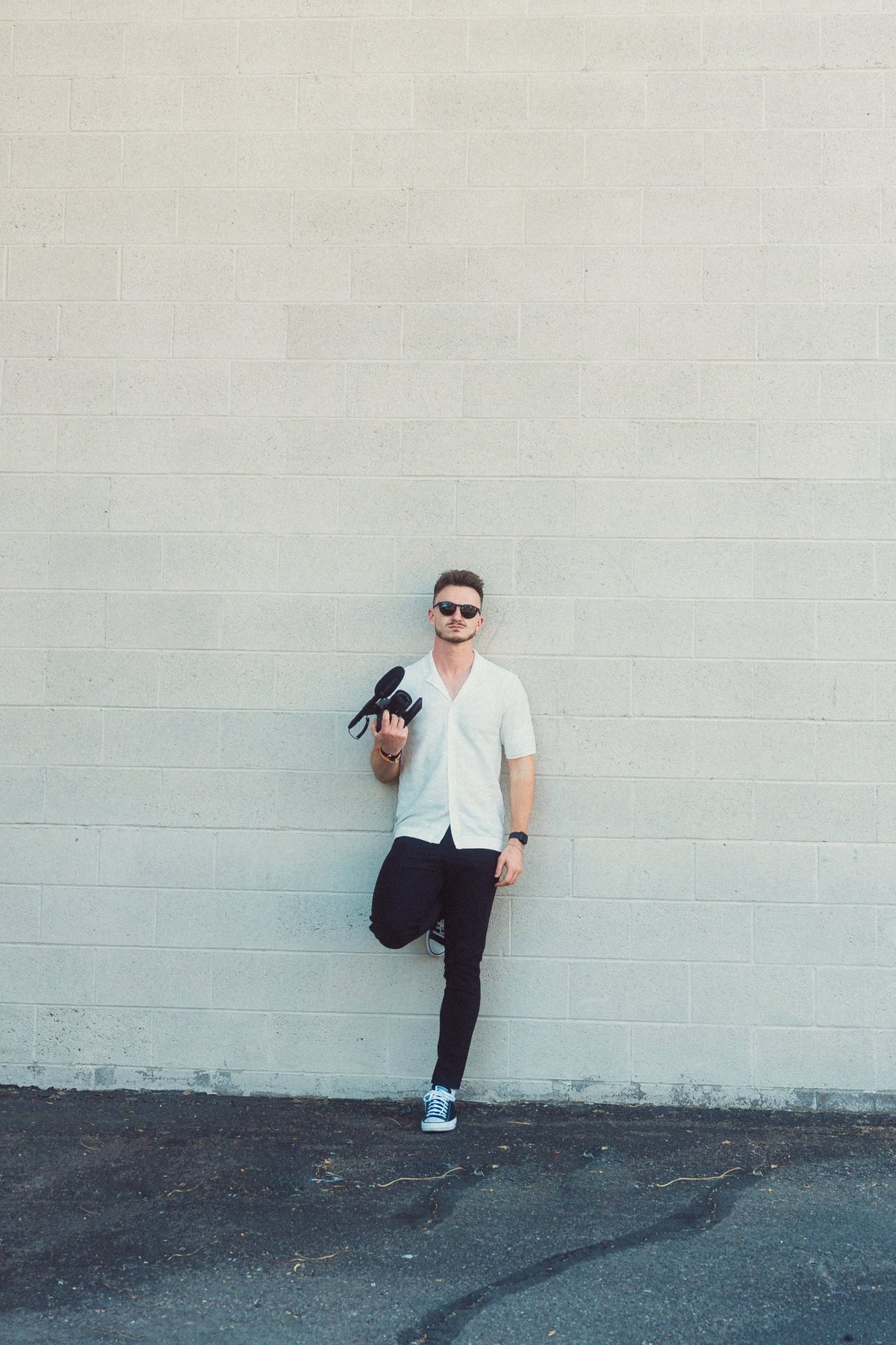Ben Radley leaning against a pale brick wall, holding a camera
